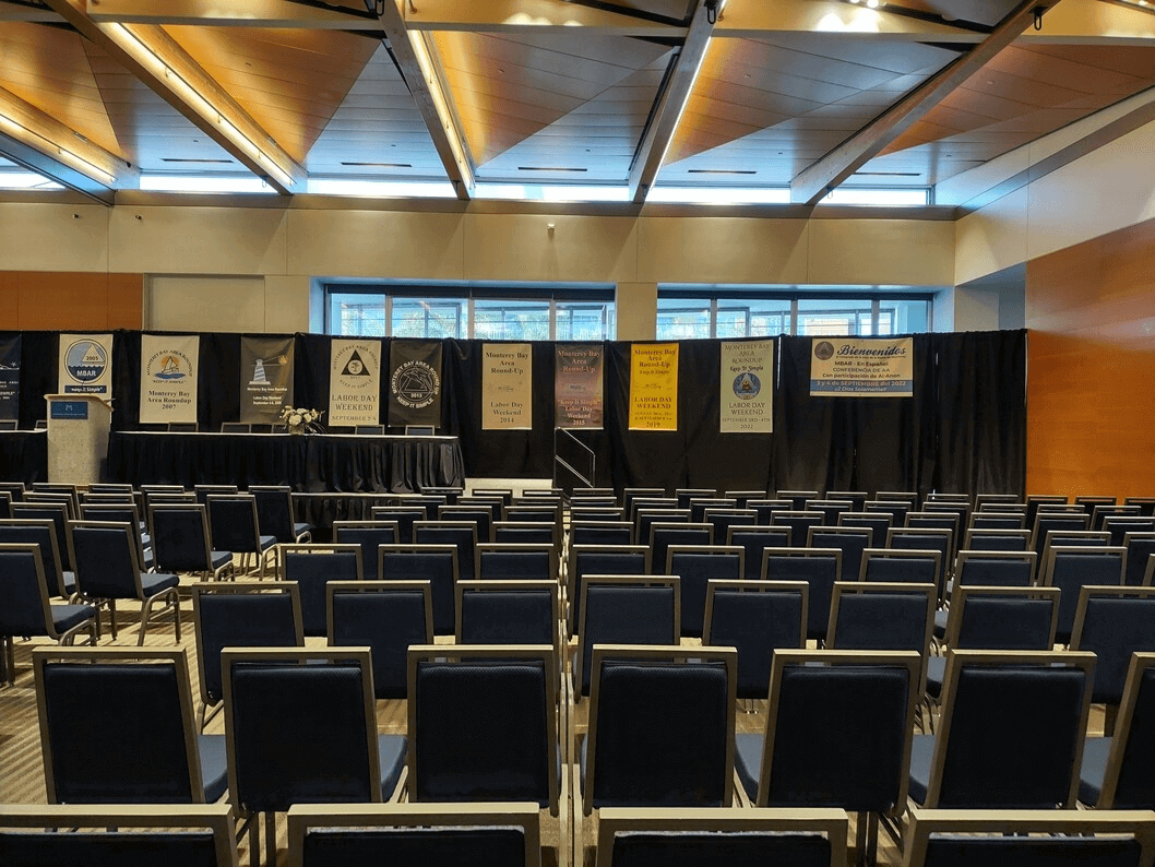 Sala de reuniones del Monterey Conference Center, sillas alineadas frente a un escenario y más allá de A.A. pancartas de conferencias.